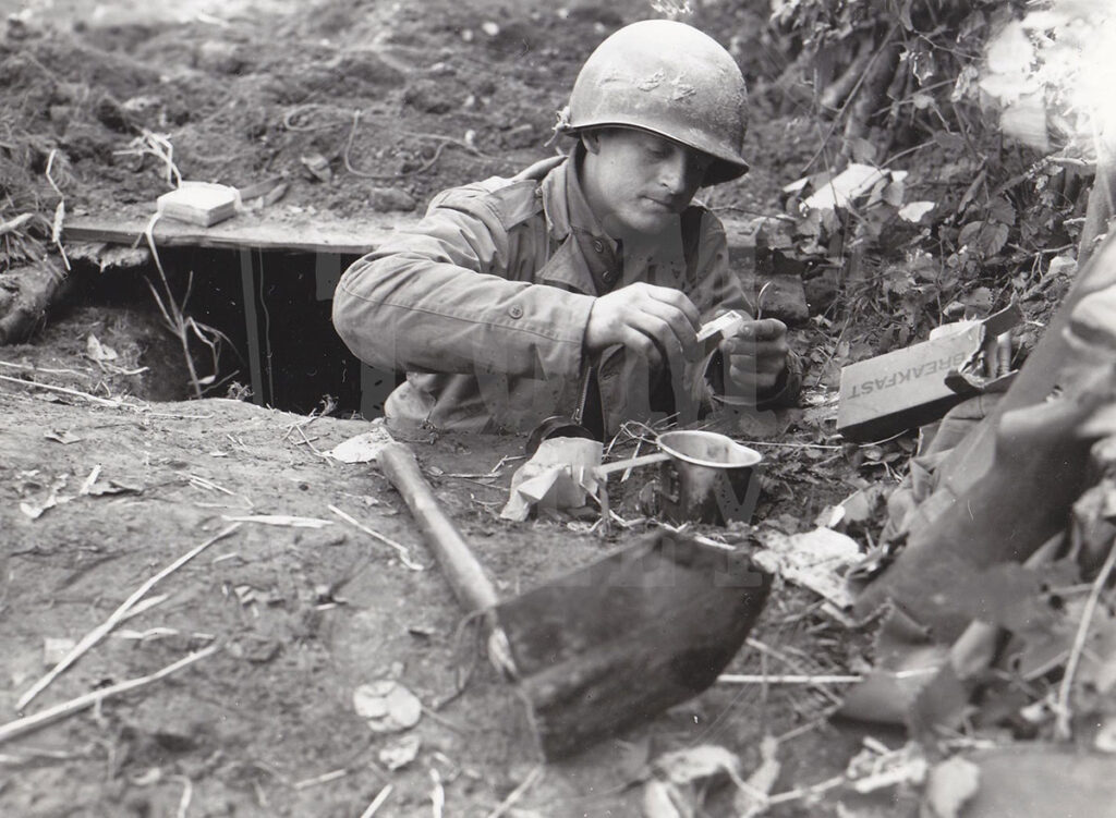 The black and white photo depicts a World War II soldier in a helmet, working with equipment in a dugout, surrounded by personal items and a shovel, and making coffee.