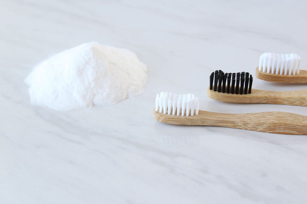 The image features a heap of baking soda alongside three bamboo toothbrushes with black and white bristles on a marble countertop.